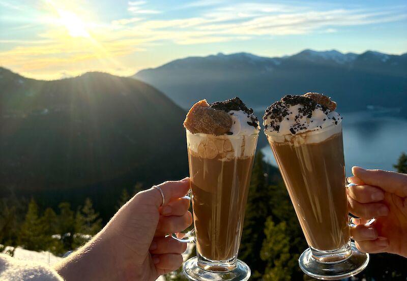 Two hands holding glasses of hot chocolate with a backdrop of the view